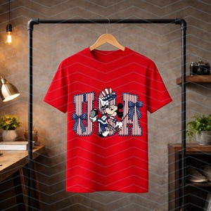 May include: A bright red short-sleeved t-shirt featuring a large graphic of Mickey Mouse wearing a patriotic hat. The letters U, S, and A are printed in a red and white striped pattern with blue star accents and blue bows on the sides.