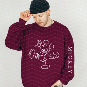 Puede incluir: Una sudadera de manga larga color granate con un dibujo de l&iacute;nea continua blanca de un rat&oacute;n de dibujos animados en la parte delantera y el texto vertical "MCKEY" en letras blancas a lo largo del lado de la manga, con un gorro de punto negro.