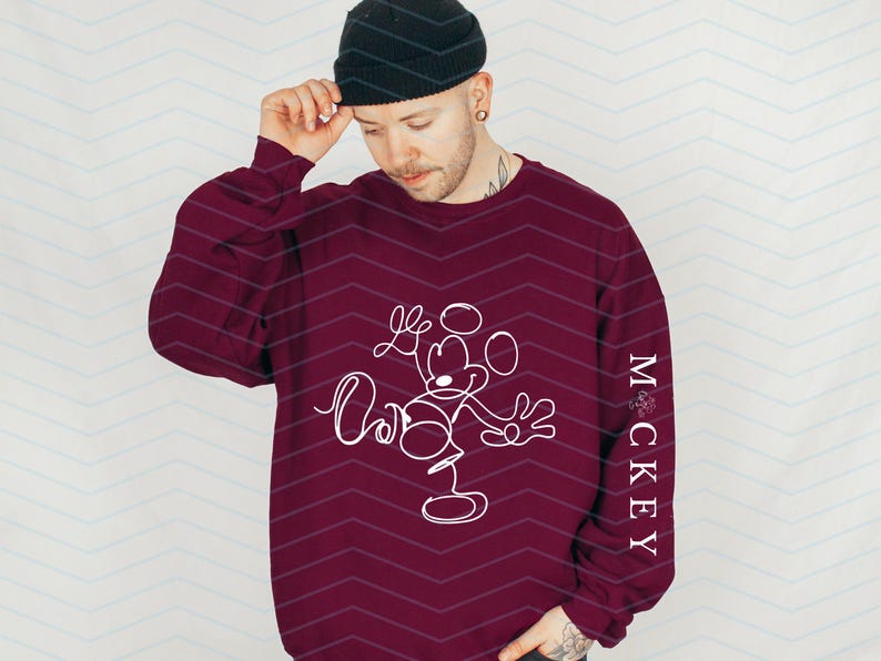 Puede incluir: Una sudadera de manga larga color granate con un dibujo de l&iacute;nea continua blanca de un rat&oacute;n de dibujos animados en la parte delantera y el texto vertical "MCKEY" en letras blancas a lo largo del lado de la manga, con un gorro de punto negro.