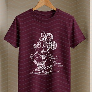 May include: A maroon t-shirt featuring a white sketch of Minnie Mouse. The design includes Minnie's bow, dress, and the text "Minnie Mouse XOXO". The shirt is hanging on a wooden hanger.