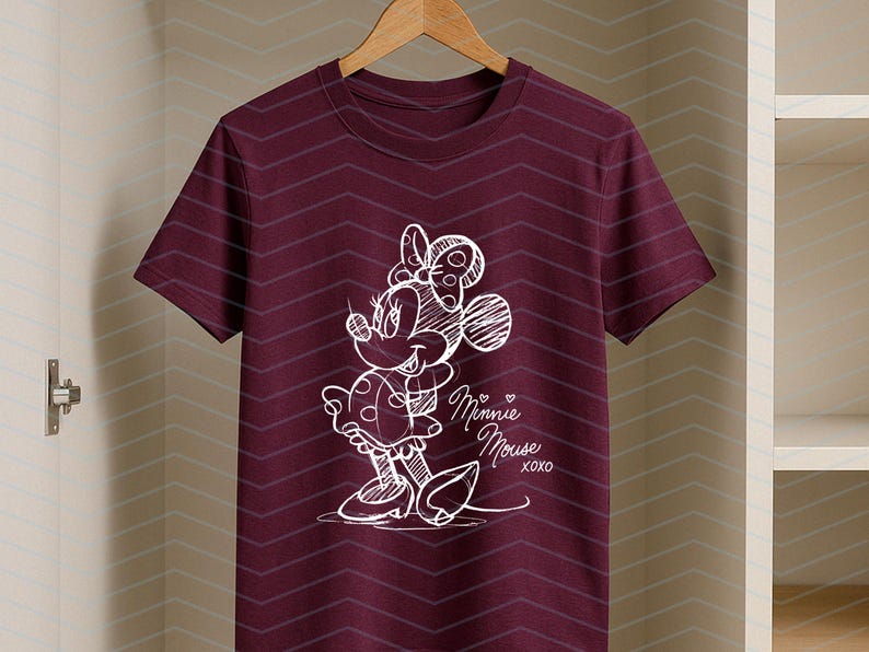 May include: A maroon t-shirt featuring a white sketch of Minnie Mouse. The design includes Minnie's bow, dress, and the text "Minnie Mouse XOXO". The shirt is hanging on a wooden hanger.