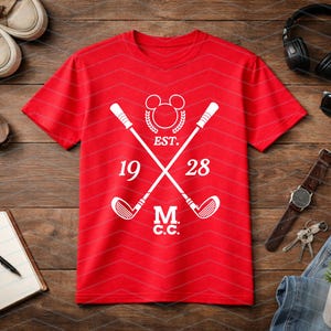 May include: Red t-shirt with a white graphic featuring crossed golf clubs, a Mickey Mouse head, and the text "EST. 1928 M.C.C.". The shirt is displayed on a wooden surface with accessories.