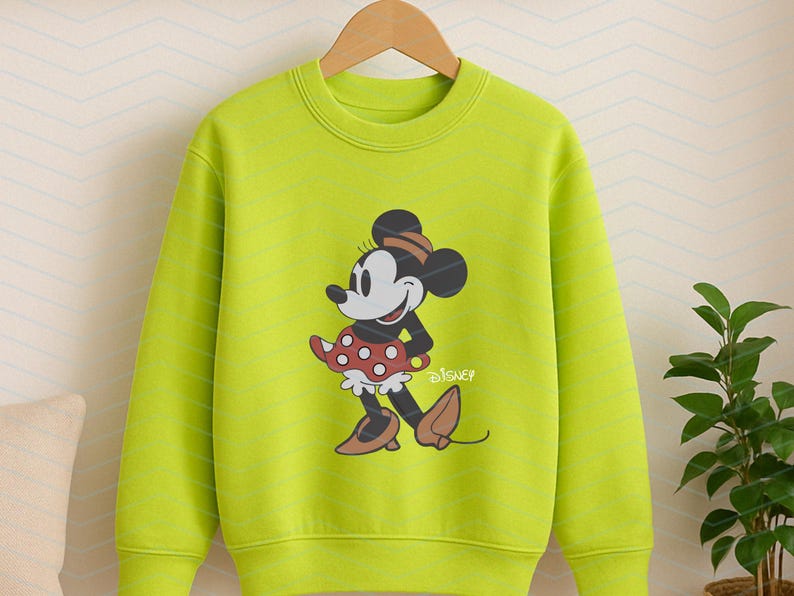 K&ouml;nnte beinhalten: Ein limettengr&uuml;nes Sweatshirt mit einer Minnie Mouse-Grafik. Minnie tr&auml;gt ihr klassisches Outfit, einen roten Polka-Dot-Rock, braune Schuhe und einen passenden Hut. Das Disney-Logo ist darunter gedruckt.