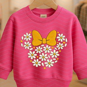 Puede incluir: Una sudadera rosa con cuello redondo y mangas largas. La parte delantera presenta un dise&ntilde;o bordado de un lazo amarillo y margaritas blancas dispuestas en forma de cabeza de Mickey Mouse. La marca "slash" est&aacute; en la etiqueta.