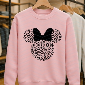May include: A light pink sweatshirt featuring a black bow and a Minnie Mouse head design. The head is filled with a leopard print pattern. The sweatshirt is hanging on a wooden hanger.