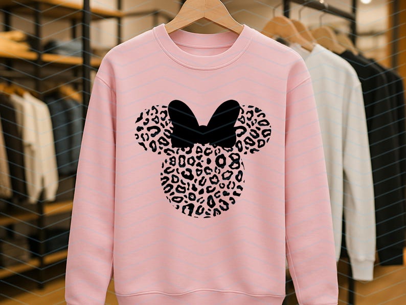 May include: A light pink sweatshirt featuring a black bow and a Minnie Mouse head design. The head is filled with a leopard print pattern. The sweatshirt is hanging on a wooden hanger.