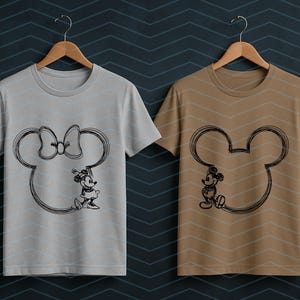 Puede incluir: Dos camisetas colgadas en perchas de madera. Una es gris claro y la otra es marr&oacute;n. Cada camiseta presenta un boceto negro de una cabeza de Mickey Mouse con un personaje en su interior. La camiseta gris claro tiene a Minnie Mouse y la camiseta marr&oacute;n tiene a Mickey Mouse.