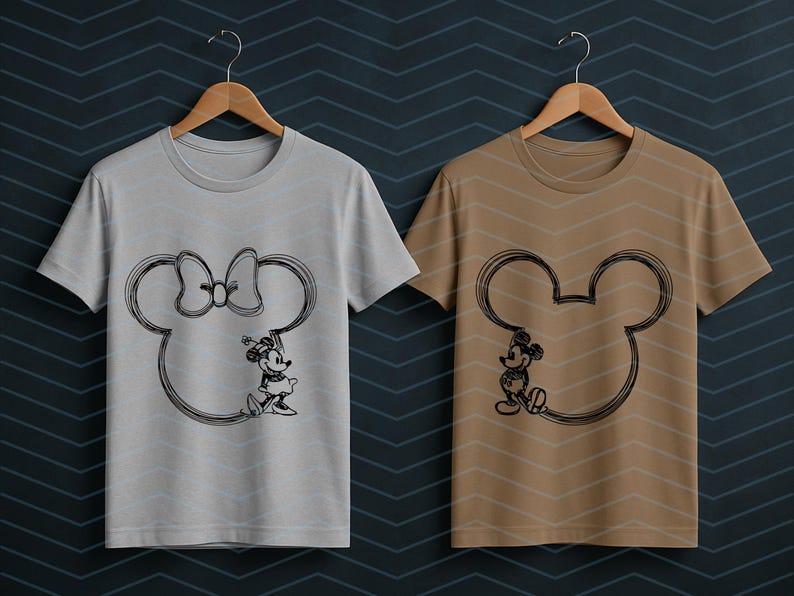 Puede incluir: Dos camisetas colgadas en perchas de madera. Una es gris claro y la otra es marr&oacute;n. Cada camiseta presenta un boceto negro de una cabeza de Mickey Mouse con un personaje en su interior. La camiseta gris claro tiene a Minnie Mouse y la camiseta marr&oacute;n tiene a Mickey Mouse.