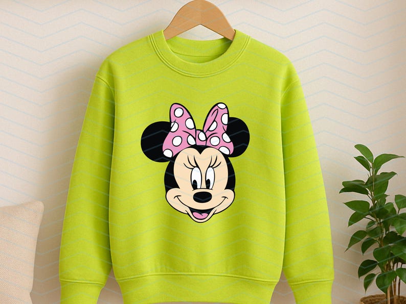 May include: A lime green sweatshirt featuring a Minnie Mouse graphic. The graphic shows Minnie's face with black ears and a pink bow with white polka dots. The sweatshirt is hanging on a wooden hanger.