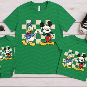 May include: Green t-shirts and a onesie featuring Donald Duck and Mickey Mouse. The shirts have a checkered background and are paired with black sneakers and sunglasses. A family matching outfit.