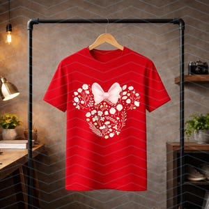 May include: A red t-shirt featuring a floral design in the shape of a mouse head with a pink bow. The design is composed of white flowers and green leaves. The t-shirt is hanging on a wooden hanger.