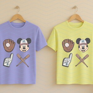May include: Two t-shirts with a purple and yellow color scheme. Both shirts feature a cartoon Mickey Mouse wearing a baseball cap with the letter "M" on it. The shirts also feature a baseball glove, a baseball, and two crossed baseball bats.