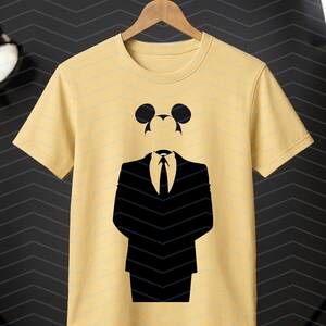 May include: A pale yellow short-sleeved t-shirt featuring a black graphic print of a suit jacket, tie, and shirt collar, topped with a silhouette of mouse ears. The design is centered on the chest of the garment, which hangs on a wooden hanger.