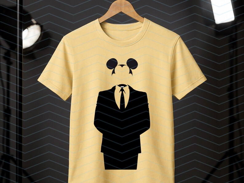 May include: A pale yellow short-sleeved t-shirt featuring a black graphic print of a suit jacket, tie, and shirt collar, topped with a silhouette of mouse ears. The design is centered on the chest of the garment, which hangs on a wooden hanger.