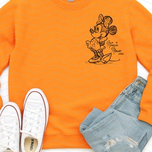 May include: An orange sweatshirt featuring a sketch of Minnie Mouse with the text "Minnie Mouse xoxo". The image also includes white sneakers and blue jeans.