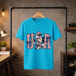May include: A bright turquoise short-sleeve t-shirt featuring a graphic print of the letters USA in a red and white striped pattern. A cartoon mouse character wearing a patriotic hat and outfit is positioned in the centre with blue bows.