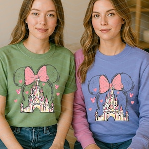 Pu&ograve; includere: Due donne indossano magliette verdi e viola con una grafica di un fiocco rosa e un castello. La grafica &egrave; un disegno stilizzato di un castello con un fiocco rosa in cima.