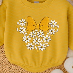 Puede incluir: Body de beb&eacute; amarillo mostaza con dise&ntilde;o de lazo y margaritas. El dise&ntilde;o presenta un lazo amarillo y margaritas blancas con centros amarillos dispuestas en forma de cabeza de Mickey Mouse. El body tiene mangas largas.