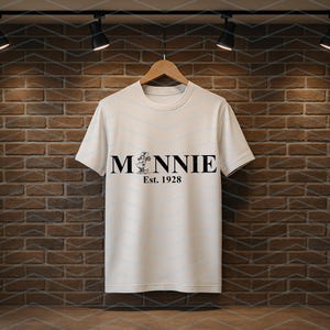 Pu&ograve; includere: Una maglietta bianca con la scritta "MINNIE" in nero, con una grafica di Minnie Mouse e il testo "Est. 1928" sotto. La maglietta &egrave; appesa a una gruccia di legno contro un muro di mattoni.
