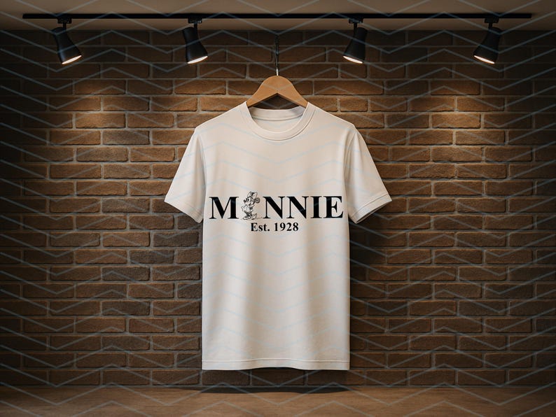 Pu&ograve; includere: Una maglietta bianca con la scritta "MINNIE" in nero, con una grafica di Minnie Mouse e il testo "Est. 1928" sotto. La maglietta &egrave; appesa a una gruccia di legno contro un muro di mattoni.