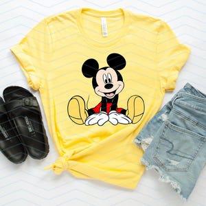 May include: A yellow t-shirt featuring a cartoon image of Mickey Mouse sitting down. The t-shirt is paired with denim shorts and black sandals.