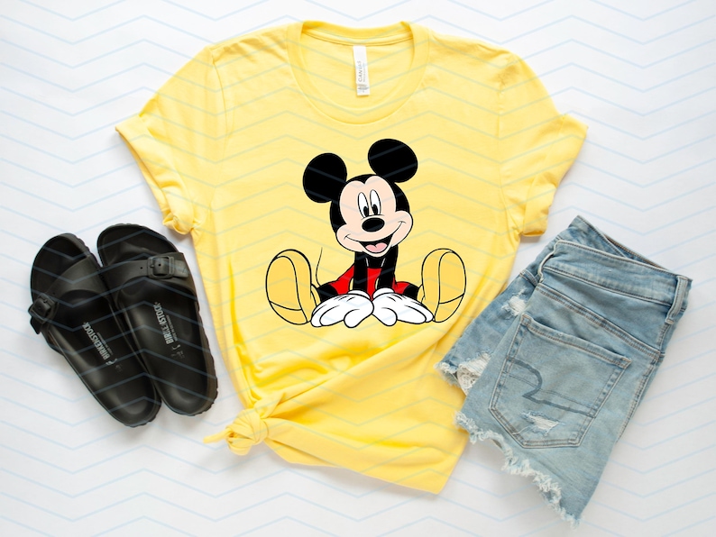 May include: A yellow t-shirt featuring a cartoon image of Mickey Mouse sitting down. The t-shirt is paired with denim shorts and black sandals.