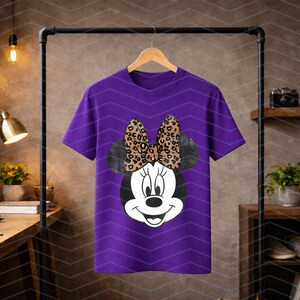 May include: A purple t-shirt featuring a Minnie Mouse graphic. The graphic includes a black and white Minnie Mouse face with a leopard print bow. The t-shirt is hanging on a wooden hanger.