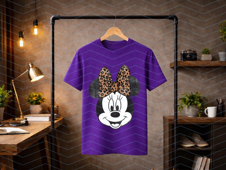 May include: A purple t-shirt featuring a Minnie Mouse graphic. The graphic includes a black and white Minnie Mouse face with a leopard print bow. The t-shirt is hanging on a wooden hanger.