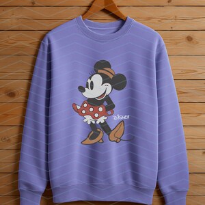 K&ouml;nnte beinhalten: Ein lila Sweatshirt mit einem Minnie Mouse-Motiv. Die Zeichentrickfigur ist mit einem roten Polka-Dot-Rock, einem schwarzen Oberteil und braunen Schuhen dargestellt. Das Disney-Logo ist darunter gedruckt.