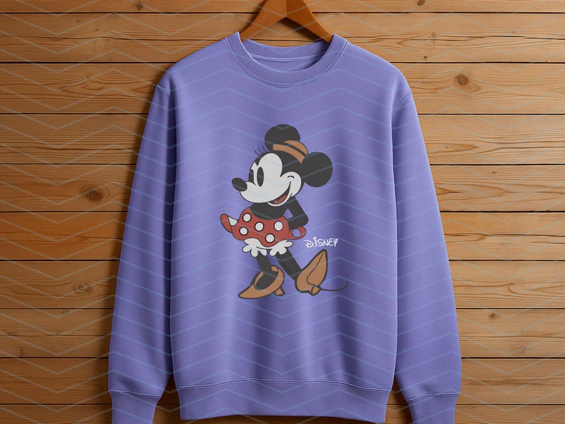 K&ouml;nnte beinhalten: Ein lila Sweatshirt mit einem Minnie Mouse-Motiv. Die Zeichentrickfigur ist mit einem roten Polka-Dot-Rock, einem schwarzen Oberteil und braunen Schuhen dargestellt. Das Disney-Logo ist darunter gedruckt.