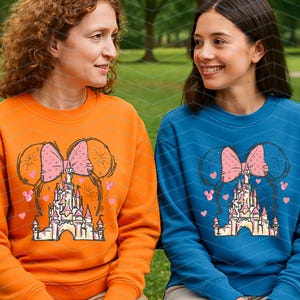 Pu&ograve; includere: Due persone indossano felpe arancioni e blu con un fiocco rosa e una grafica di un castello. La grafica presenta un fiocco rosa con un castello al centro e cuori rosa e fuochi d'artificio intorno.