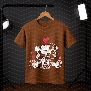 Peut inclure: Un t-shirt marron avec un dessin blanc de Mickey et Minnie Mouse s'embrassant, avec des coeurs roses. Le dessin est centr&eacute; sur le t-shirt. Le t-shirt est suspendu &agrave; un cintre en bois.