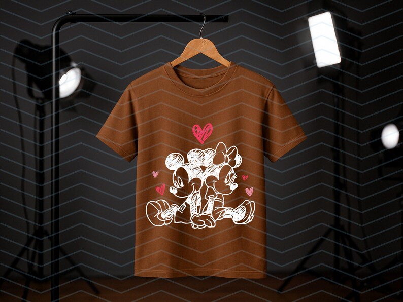Peut inclure: Un t-shirt marron avec un dessin blanc de Mickey et Minnie Mouse s'embrassant, avec des coeurs roses. Le dessin est centr&eacute; sur le t-shirt. Le t-shirt est suspendu &agrave; un cintre en bois.
