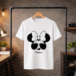 May include: White t-shirt featuring a black and white Minnie Mouse design. The design includes sunglasses, a bow, and the name "Isabella" in a script font. The shirt is hanging on a wooden hanger.