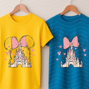 Pu&ograve; includere: Due t-shirt con un design grafico di un fiocco rosa e un castello. Il castello &egrave; rosa e bianco con una porta gialla. Il fiocco &egrave; sopra il castello. Le t-shirt sono gialla e blu.