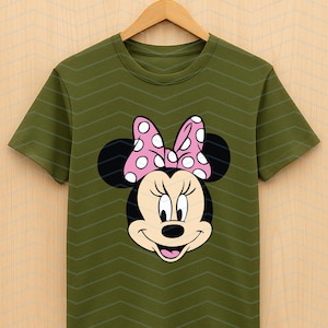 May include: Olive green t-shirt featuring a Minnie Mouse graphic. The graphic shows Minnie's face with black ears, a pink bow with white polka dots, and a smiling expression. The shirt is hanging on a wooden hanger.