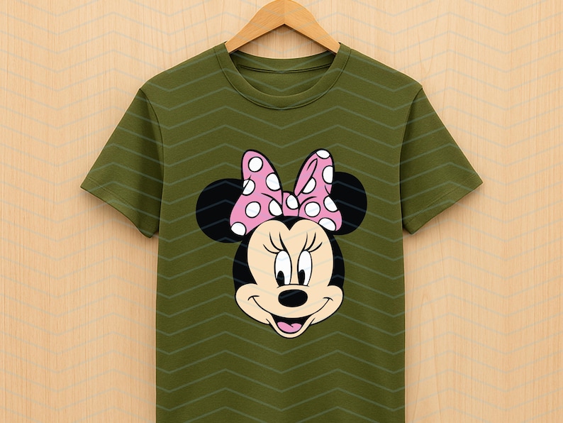 May include: Olive green t-shirt featuring a Minnie Mouse graphic. The graphic shows Minnie's face with black ears, a pink bow with white polka dots, and a smiling expression. The shirt is hanging on a wooden hanger.