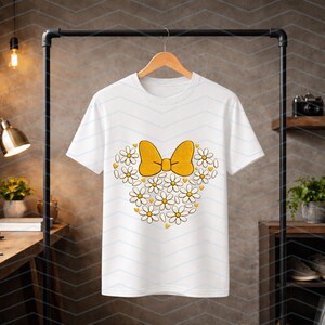 Puede incluir: Camiseta blanca con un lazo amarillo y un dise&ntilde;o en forma de coraz&oacute;n hecho de margaritas blancas con centros amarillos. La camiseta est&aacute; colgada en una percha negra.