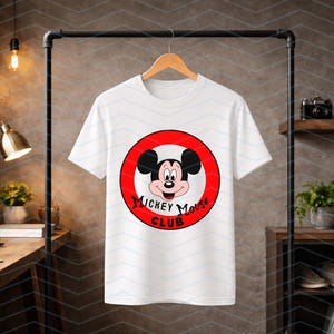 May include: A white short-sleeved t-shirt featuring a circular graphic on the chest. The graphic displays the face of Mickey Mouse inside a red ring with the black text Mickey Mouse Club printed in a stylized font along the bottom edge.