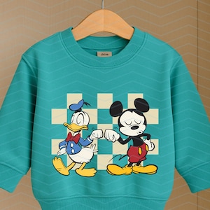 May include: Teal-colored sweatshirt featuring Donald Duck and Mickey Mouse bumping fists. The cartoon characters are set against a checkered background. The long-sleeved top has a crew neck and is hanging on a wooden hanger.