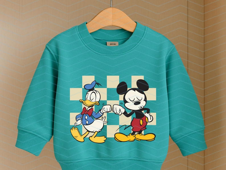 May include: Teal-colored sweatshirt featuring Donald Duck and Mickey Mouse bumping fists. The cartoon characters are set against a checkered background. The long-sleeved top has a crew neck and is hanging on a wooden hanger.
