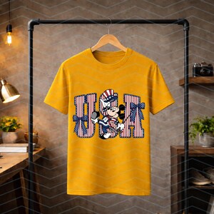 May include: A mustard yellow short-sleeve t-shirt featuring a graphic print of Mickey Mouse wearing a patriotic top hat. The character is positioned in front of large, block-style letters spelling USA, which are decorated with red and white stripes.