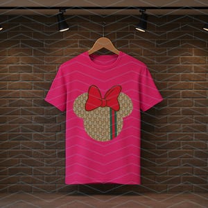 Puede incluir: Una camiseta de manga corta de color rosa brillante con un gr&aacute;fico central de la silueta de una cabeza de rat&oacute;n. La silueta tiene un patr&oacute;n repetitivo en beige y marr&oacute;n con una franja vertical verde y roja, rematada con un gran lazo rojo.