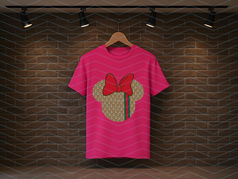 Puede incluir: Una camiseta de manga corta de color rosa brillante con un gr&aacute;fico central de la silueta de una cabeza de rat&oacute;n. La silueta tiene un patr&oacute;n repetitivo en beige y marr&oacute;n con una franja vertical verde y roja, rematada con un gran lazo rojo.