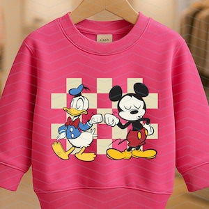 May include: A bright pink sweatshirt featuring a graphic of Donald Duck and Mickey Mouse bumping fists. The cartoon characters are set against a checkered background. The sweatshirt has long sleeves and a crew neck.