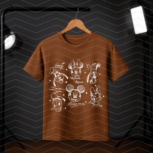 May include: A brown t-shirt featuring white sketches of Disney characters including Mickey Mouse, Minnie Mouse, Donald Duck, Daisy Duck, Goofy, and Pluto. The names of the characters are written below each sketch. The shirt is hanging on a wooden hanger.