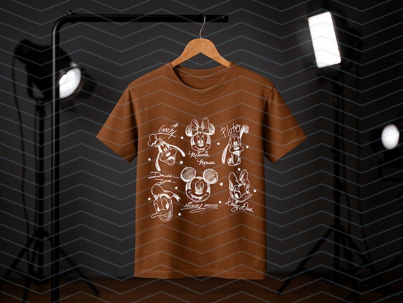 May include: A brown t-shirt featuring white sketches of Disney characters including Mickey Mouse, Minnie Mouse, Donald Duck, Daisy Duck, Goofy, and Pluto. The names of the characters are written below each sketch. The shirt is hanging on a wooden hanger.