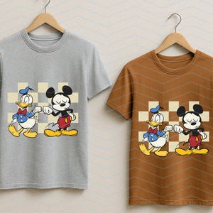 May include: Two t-shirts, one gray and one brown, each featuring a cartoon graphic of Mickey Mouse and Donald Duck bumping fists. The characters are set against a checkered background. The shirts are hanging on wooden hangers.