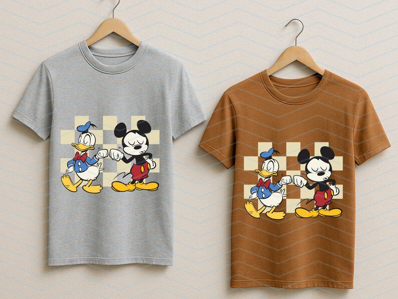 May include: Two t-shirts, one gray and one brown, each featuring a cartoon graphic of Mickey Mouse and Donald Duck bumping fists. The characters are set against a checkered background. The shirts are hanging on wooden hangers.