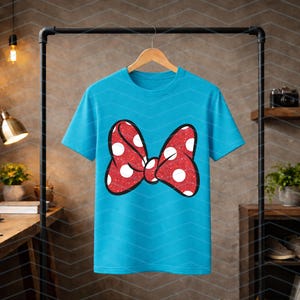 Puede incluir: Una camiseta turquesa con un gran lazo rojo con lunares blancos. El lazo tiene una textura brillante y un contorno negro. La camiseta est&aacute; colgada en una percha de madera sobre un fondo neutro.